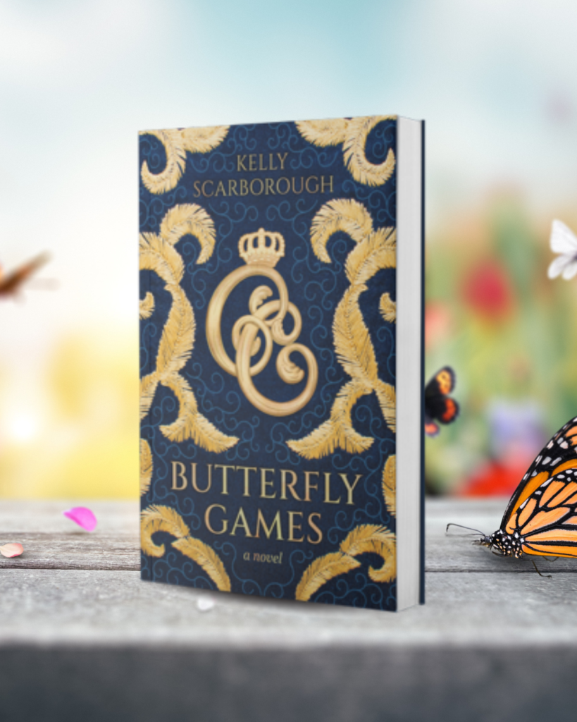 A copy of Butterfly Games by Kelly Scarborough stands upright on a wooden surface, its dark blue and gold embroidered-style cover in focus. Monarch butterflies and soft, colorful flowers appear around the book against a bright, blurred background.